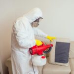 pest control worker in uniform spraying pesticides under couch in living lounge room